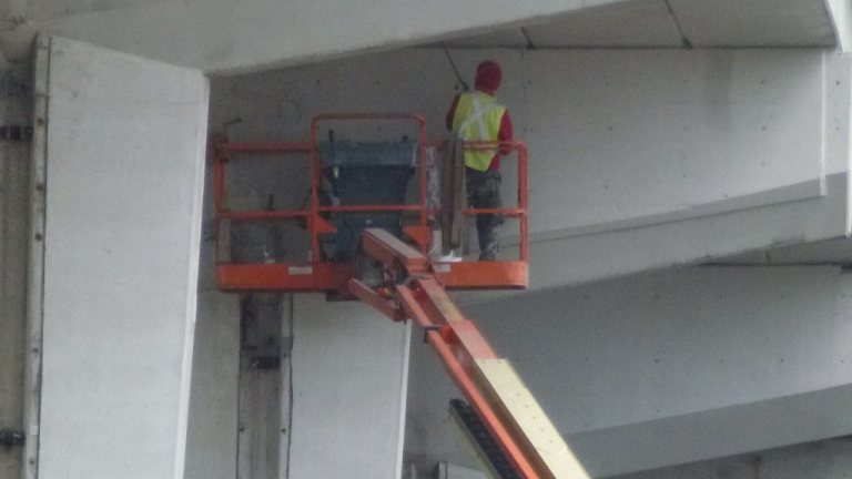 Worker in a hard hat and harness in the basket of a boom lift under a concrete expressway