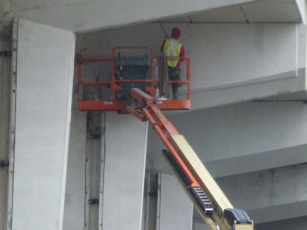 Worker in a hard hat and harness in the basket of a boom lift under a concrete expressway