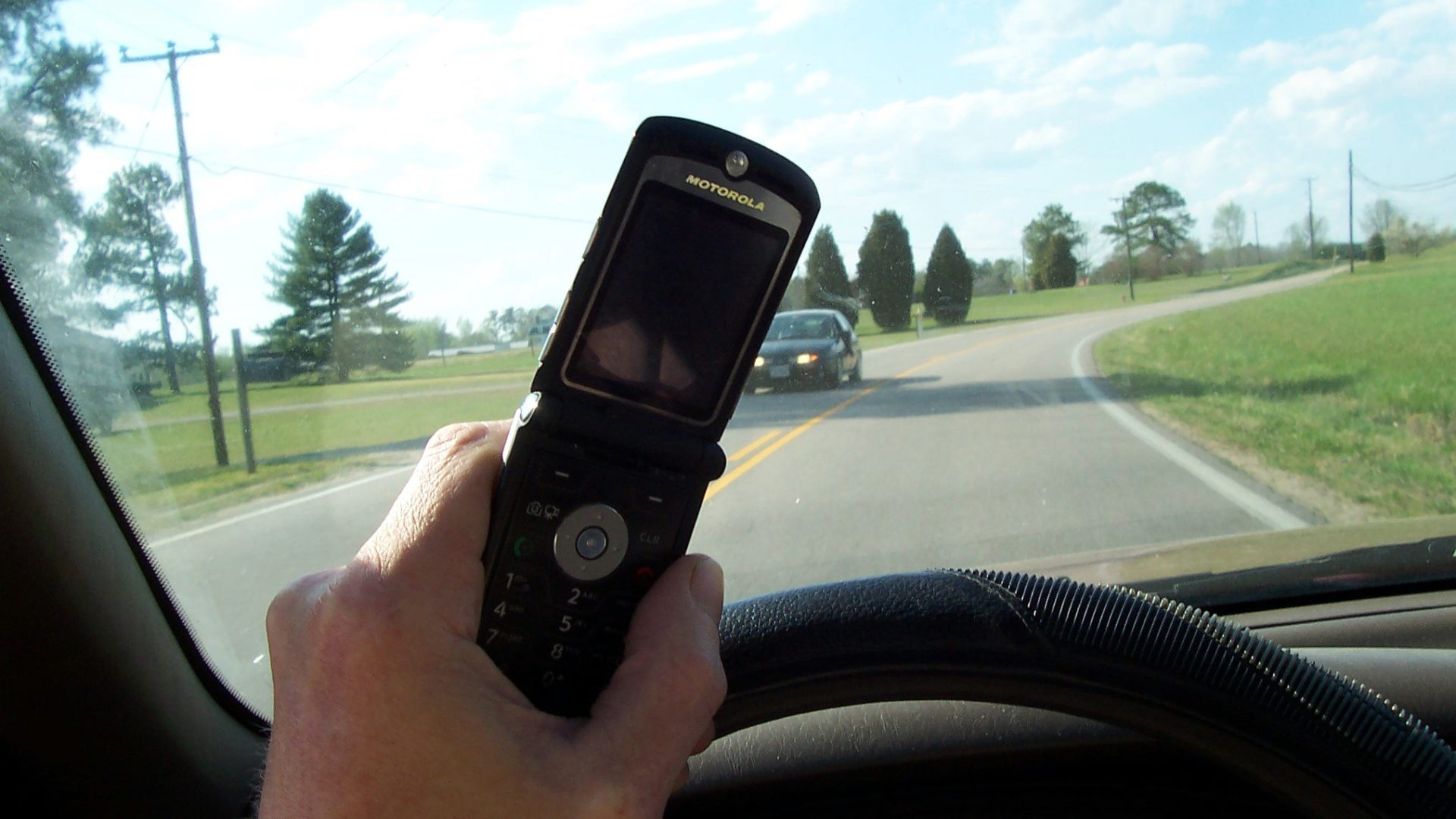 Driver holding a cell phone over the steering wheel on a two-lane country road