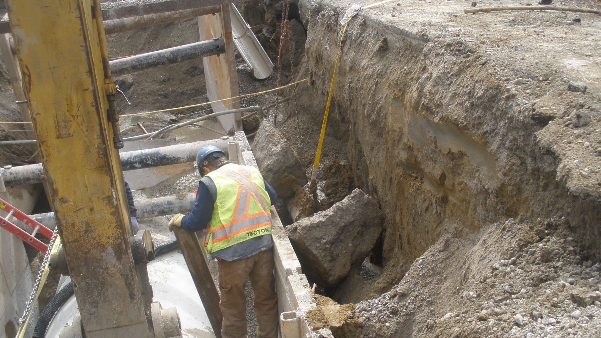 Worker in a high-visibility vest working beside a shored trench at a city sewer job